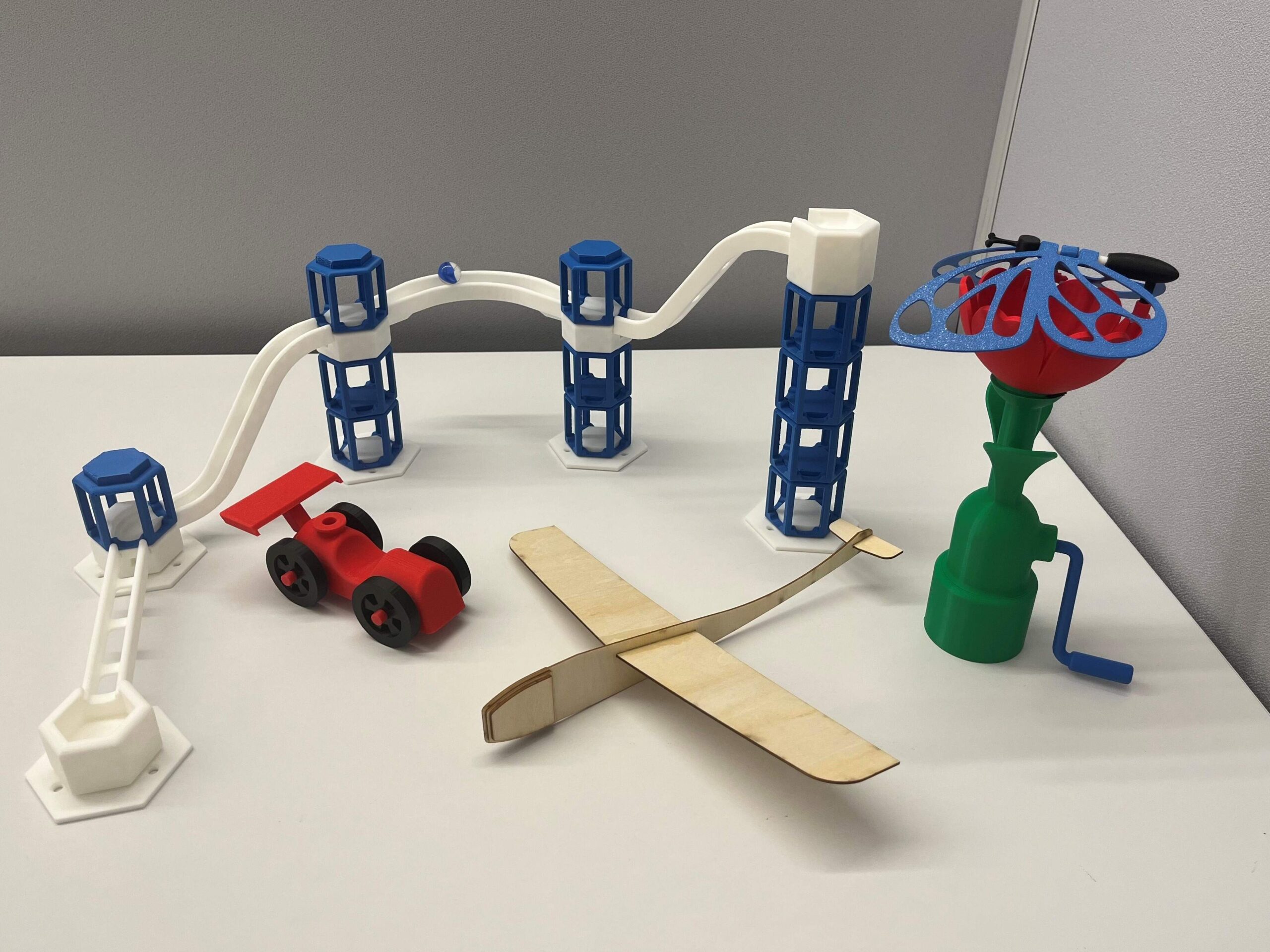 A collection of models, designed to be educational training materials to teach students to use xDesign, sit on a table.  Pictured is a marble run, a balloon-powered racer, a balsa-wood glider, and a kinetic sculpture.
