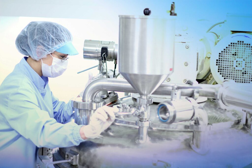 A worker in PPE sits in front of a complex machine in a factory setting - virtual twins in pharma - Dassault Systemes blog 